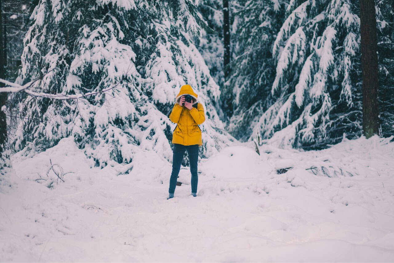 Photographer in yellow jacket taking winter landscape photo in snow-covered forest.