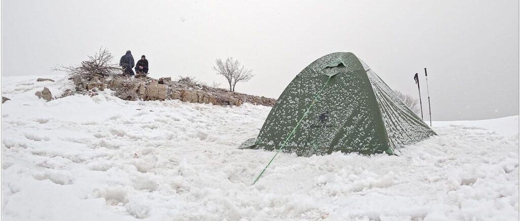 Warm winter survival tent in snow with trekking poles and snow-covered landscape.