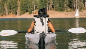 Light gray kayaking backpack on woman paddling on a calm lake surrounded by trees.