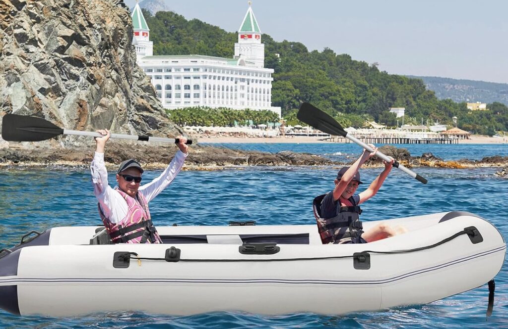 Inflatable kayak with two people paddling on clear blue water near rocky shoreline and scenic background.
