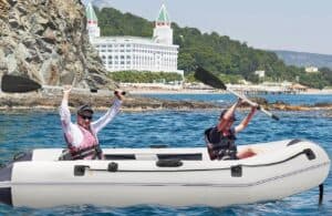 Inflatable kayak with two people paddling on clear blue water near rocky shoreline and scenic background.