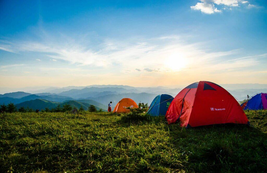 Colorful camping tents set up on a grassy mountain hilltop at sunset.